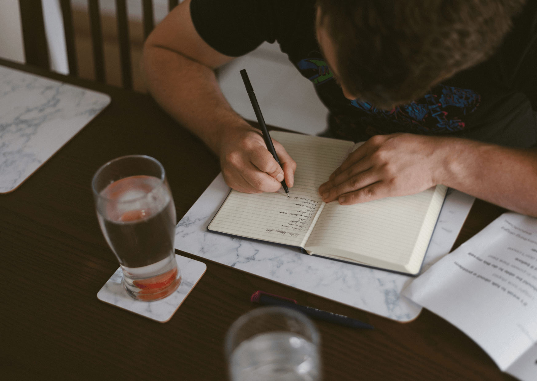 Person writing in a notebook with a glass of water on a table