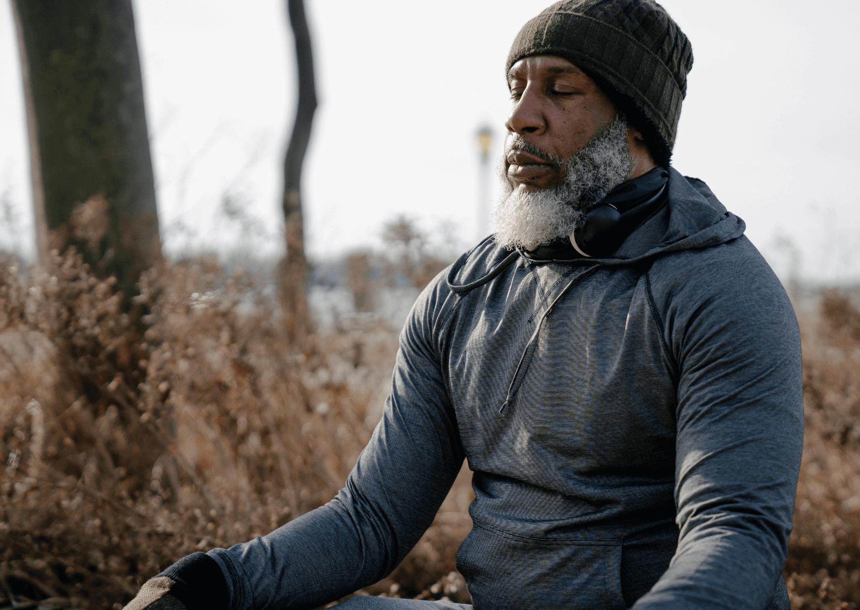 Man sitting outdoors in a forested area wearing a dark hoodie and beanie.